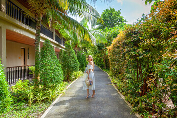 Fototapeta na wymiar Young woman in white dress strolling through lush green trees in tropical resort. Concept of summer vacation and relaxation.