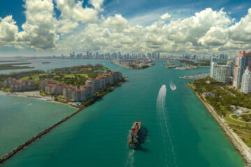 Naklejka premium Commercial container ship entering Miami port harbor through main channel near South Beach. Luxurious hotels and residential buildings on waterfront and high skyscraper towers of downtown in distance