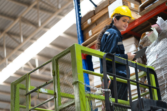 Caucasian businesswoman workers using clipboard checking stock while standing on scissor lift in warehouse	