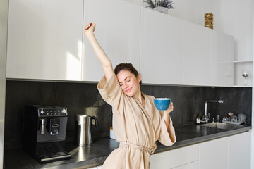 Portrait of happy girl dancing with coffee in the kitchen, wearing bathrobe, enjoying her morning...