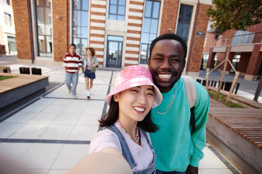 POV of two ethnic young students taking selfie photo together on college campus and smiling at camera happily