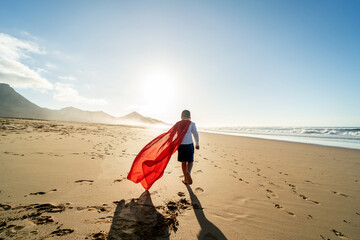 Superhero child walking on the sandy, sunset beach, wearing red cape. Back view. Full length....