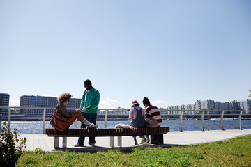 Back view at diverse group of young people relaxing outdoors by river against city skyline, copy...