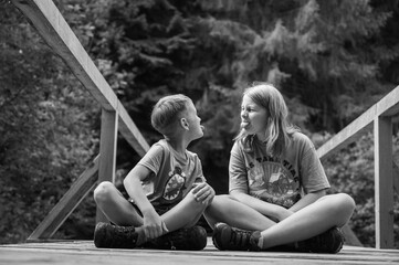 Black and white, small cute and emotional children boy and girl sit on wooden surface, bridge in forest and look straight. Ukrainian Carpathians, recreation and travel, entertainment