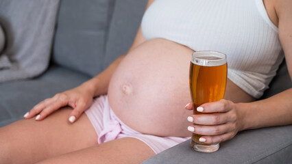 Close-up of the belly of a pregnant woman holding a glass of beer while sitting on the sofa.