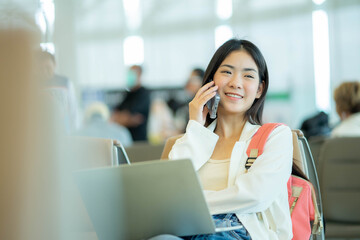 Happy Young Asian woman sitting at the airport terminal departure lounge and talking on the cell phone while waiting for a flight. Travel concept. - obrazy, fototapety, plakaty