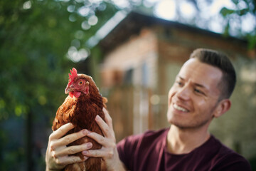 Portrait of smiling farmer while standing outside henhouse and holding hen on organic farm. . © Chalabala