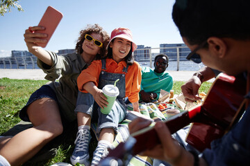 Multiethnic group of young people taking selfies outdoors enjoying picnic on grass in sunlight, copy space