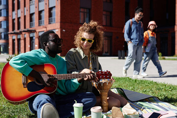 Portrait of gen Z young couple enjoying picnic on grass in city and playing guitar, copy space