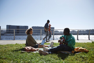 Side view at group of young people enjoying picnic outdoors at riverside park