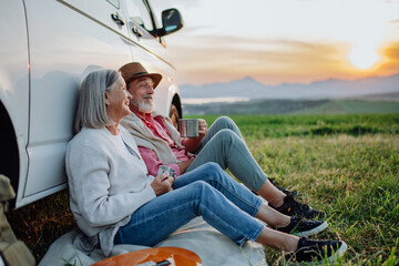 Senior couple sitting by car and drinking coffee after long drive during their roadtrip. Elderly...