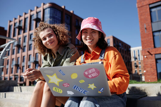 Colorful portrait of two young women using computer outdoors while sitting on concrete stairs in city