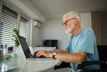 Senior man in wheelchair working from home during retirement. Elderly man using digital technologies, working on laptop. Concept of seniors and digital skills.
