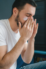 Bearded handsome man applying face cream sitting on the sofa at home. Beauty products for his face, moisturising or nourishing skin