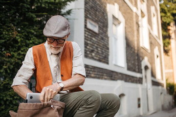Senior man sitting on street curb in the city. Portrait of elderly man searching for things in a backpack. - Powered by Adobe