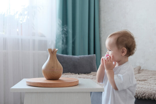 Water diffuser, wooden jug with steam. A girl of European appearance, two years old, joyful, in white clothes in the room. Blurred background. Concept of health care, air humidification
