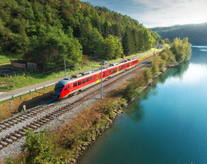 Aerial view of red modern high speed train moving near river in alpine mountains at sunset in autumn. Top view of train, rural railroad, lake, road, green trees in fall. Railway station in Slovenia - 660105373