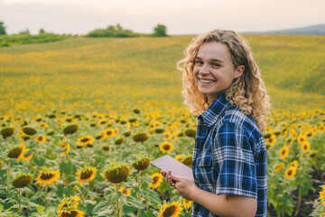 Naklejka na ściany i meble Happy woman farmer with curly hair is at sunflower field, holding smart tablet, smiling while looking at camera. Concept, agriculture occupation, using wireless