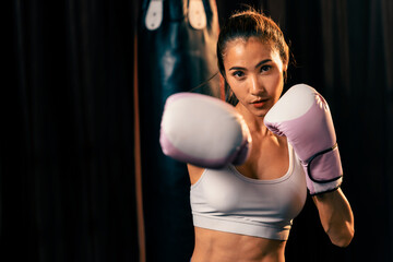 Asian female Muay Thai boxer punch fist in front of camera in ready to fight stance posing at gym with boxing equipment in background. Focused determination eyes and prepare for challenge. Impetus