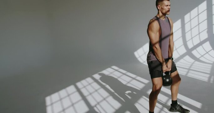 Muscular young man has strength workout at studio with white background and window shadow. Sporty male bodybuilder doing squatting with kettlebell in hands