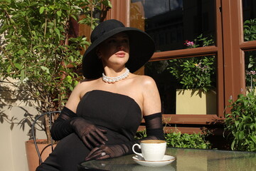 Pregnant beautiful elegant lady in a black dress and a black hat with a large pearl necklace drinks...