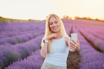 Lavender field. Pretty woman with blond hair walking between rows of lavender flowers with mobile phone, taking selfie, communicating in social networks, relaxing, resting, enjoying nature