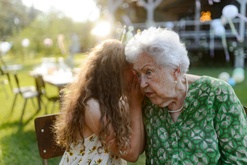 Girl whispering a secret into grandmother's ear at a garden party. Love and closeness between grandparent and grandchild.