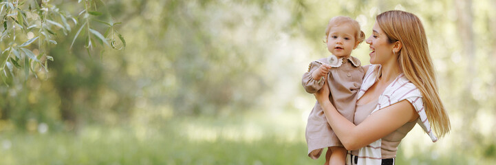 A young european woman in casual, comfortable beige clothes holding a small child in her arms in the park. Happy moments of life, family vacation in nature with copy space. Banner with copy space.