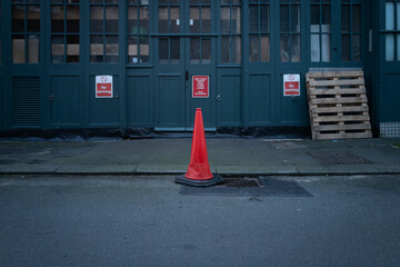 Shallow focus of a single red traffic cone seen outside a warehouse entrance on a busy street. Used...