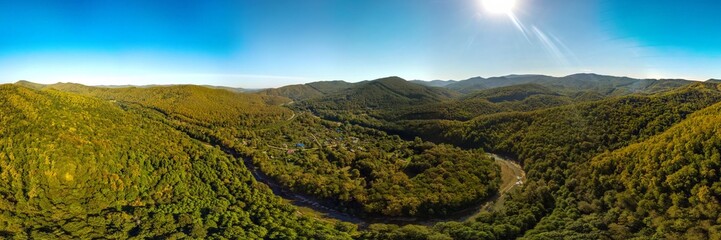 a farm near a river bend surrounded by the Western Caucasus mountains covered with green deciduous forest on a sunny day in early autumn - aerial panorama