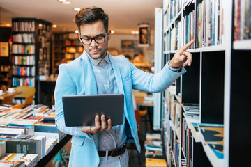 Young handsome man sitting in modern bookstore, reading and using his laptop or notebook computer.