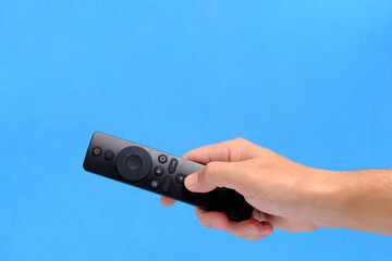 Hand blue background holds TV remote by pressing button with finger.