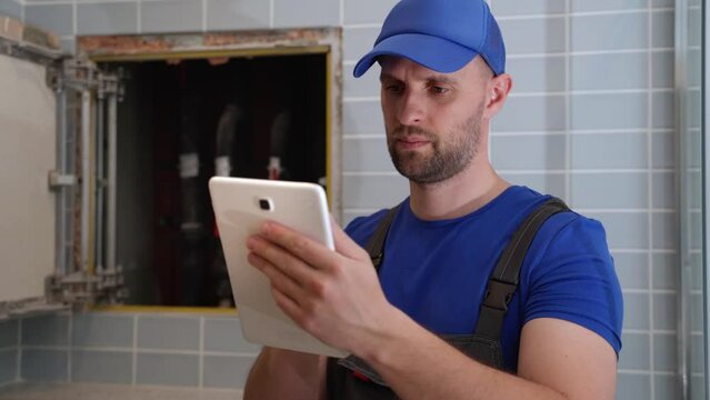 Male plumber checks pipes for central hot and cold water supply of apartment. Male plumber performs diagnostics using a tablet computer.