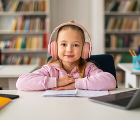 Pretty schoolgirl sitting at desk in wireless headphones, smiling