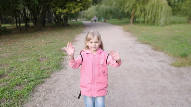 cute little girl walks through the park in a pink jacket and gestures goodbye with both hands slow motion