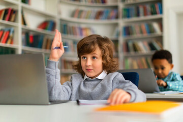 Fototapeta na wymiar Schoolboy raising hand while using laptop in class
