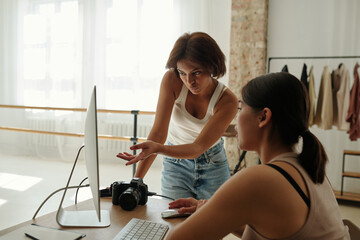 Young female fashion model pointing at photo shot on screen of computer while discussing it with professional photographer after session