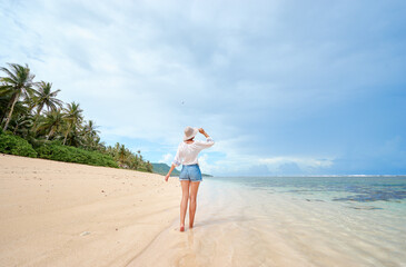 Fototapeta na wymiar Vacation on the seashore. Back view of young woman walking away on the beautiful tropical white sand beach.