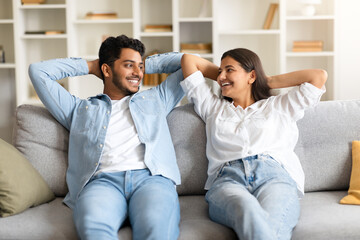 Obraz premium Happy indian couple on sofa, hands behind head, smiling at each other