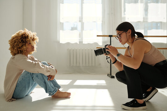 Side view of young female photographer with photocamera taking pictures of fashion model in stylish casualwear sitting on the floor by wall