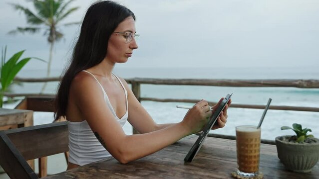 Female freelancer draws with stylus on a graphics tablet in cafe with ocean view, palm trees. Artist girl working on device by the seaside. Young illustrator on remote work day on tropical island.