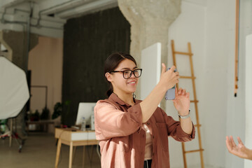 Happy young female photographer with smartphone taking pictures of fashion model while standing in spacious studio during photo session