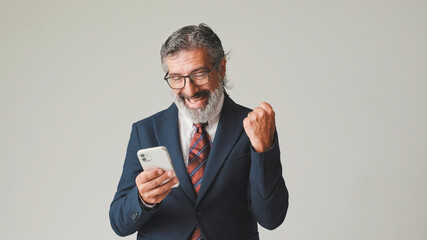 Businessman in glasses with gray hair, looking with smile at the mobile phone screen, getting good deal, winning, isolated on gray background in the studio