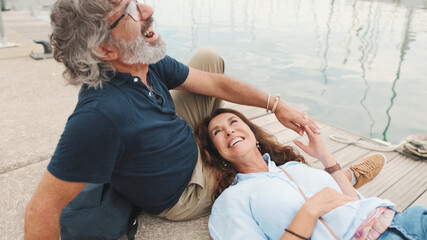 Laughing middle aged couple, retired elderly couple enjoy outdoor recreation together, relax and chat while lying on the pier