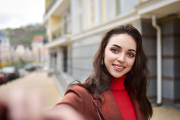 A young woman in a warm coat on the street holds her phone in her hand and makes a selfie.