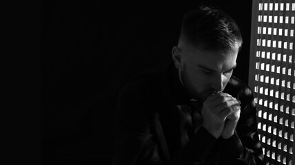 Grayscale portrait of young praying priest in confession booth