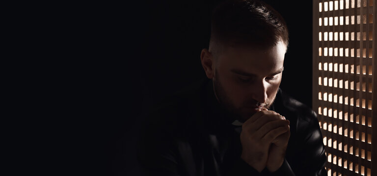 Portrait of young praying priest in confession booth