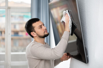 Tidy young bearded man cleaning his house, wiping dust on screen of TV in living room with rag..