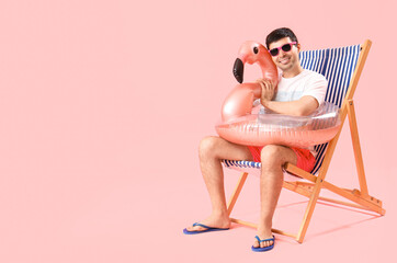 Fototapeta premium Young man with swim ring sitting in deck chair on pink background