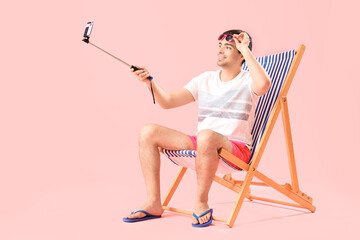 Young man taking selfie in deck chair on pink background